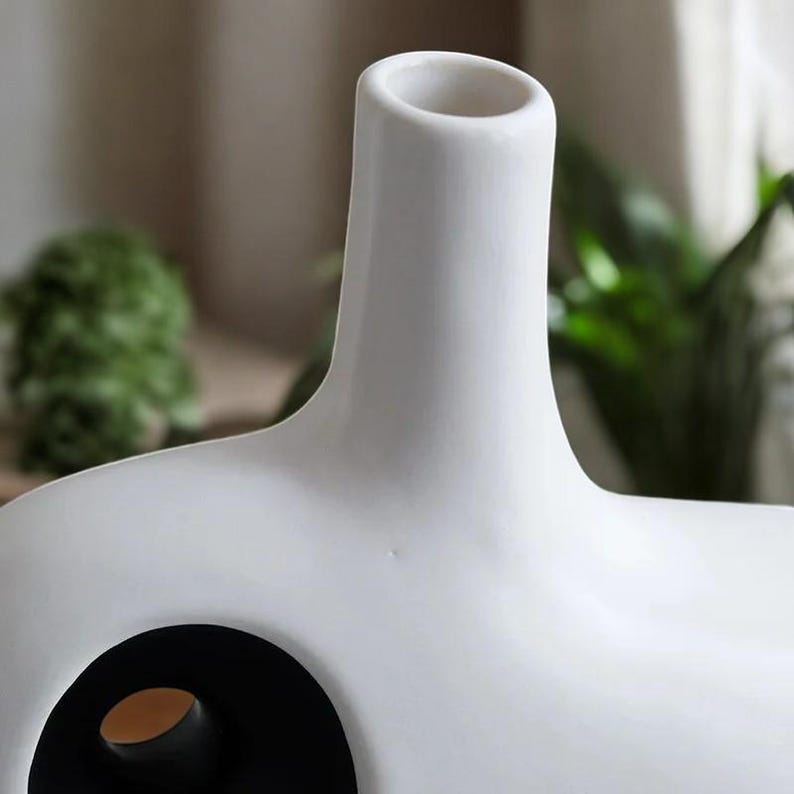 May include: A modern white ceramic vase with a unique design. The vase features a cylindrical neck and a rounded body with a large circular opening. The interior of the opening is black, creating a striking contrast. The vase is a decorative home accent.
