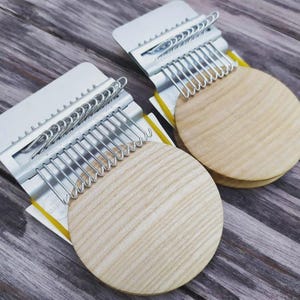 Może przedstawiać: Dwa kalimby, każdy z jasnym drewnianym pudłem rezonansowym i metalowymi zębami, są wyświetlane na drewnianej powierzchni. Kalimby mają srebrny metalowy uchwyt na zęby i okrągłą drewnianą podstawę. Tło to zwietrzała drewniana powierzchnia.