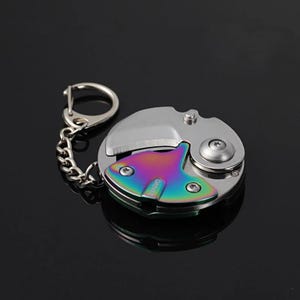 Pode incluir: Uma pequena faca de bolso redonda prateada com um detalhe colorido em arco-íris e um porta-chaves. A lâmina da faca está parcialmente aberta, revelando uma borda afiada. A palavra "DRIVE" é visível no detalhe arco-íris.