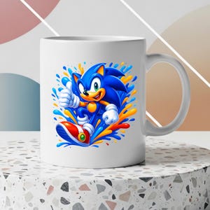 May include: A white ceramic mug featuring a vibrant graphic of the blue character Sonic giving a thumbs up. The design includes dynamic splashes of blue, yellow, and orange paint surrounding the character, who wears white gloves and red shoes.