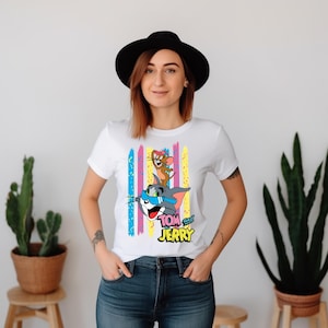 May include: A white short-sleeved t-shirt featuring a graphic of Tom and Jerry with Tom wearing blue sunglasses and Jerry wearing a pink bow. The design includes vertical colourful brush strokes in pink, yellow, and blue, with the text Tom and Jerry.