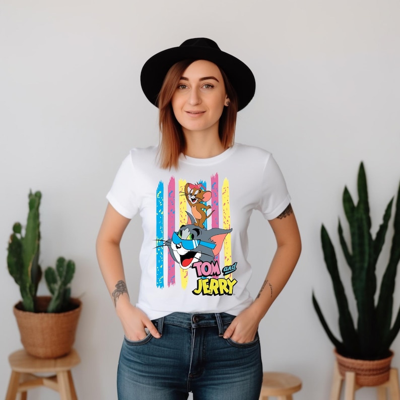 May include: A white short-sleeved t-shirt featuring a graphic of Tom and Jerry with Tom wearing blue sunglasses and Jerry wearing a pink bow. The design includes vertical colourful brush strokes in pink, yellow, and blue, with the text Tom and Jerry.