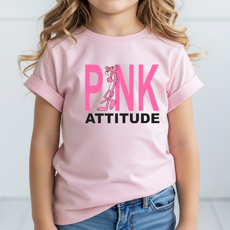 May include: A light pink short-sleeved t-shirt featuring a graphic of the Pink Panther character standing in place of the letter I in the word PINK, printed in large hot pink letters, with the word ATTITUDE printed in black text underneath.