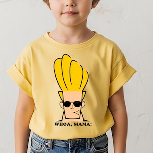 May include: A light yellow short-sleeved t-shirt featuring a graphic print of the cartoon character Johnny Bravo with his signature tall blonde hair and black sunglasses. Below the character, the text reads WHOA, MAMA! in bold black letters.