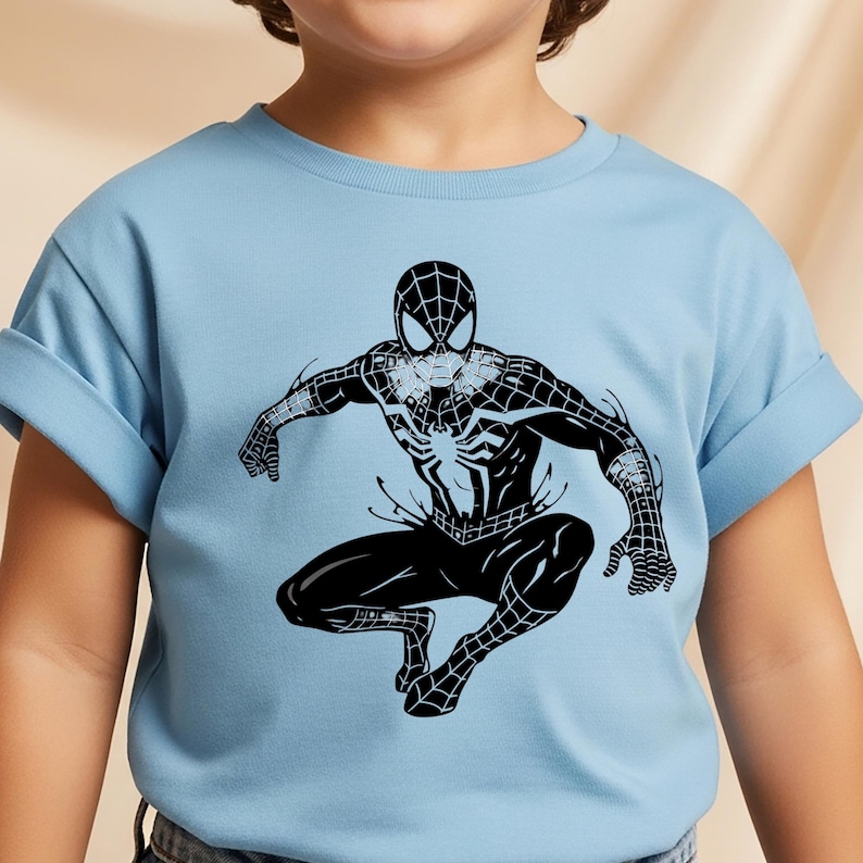 May include: A light blue short-sleeved t-shirt featuring a large, centred black graphic print of a superhero character in a web-patterned suit. The character is depicted in a dynamic crouching pose with arms extended, showcasing detailed web lines.