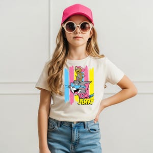 May include: A cream-coloured short-sleeve t-shirt featuring a graphic print of Tom and Jerry with blue sunglasses, set against vertical pink, blue, and yellow stripes. The shirt is paired with blue denim jeans, a bright pink baseball cap, and sunglasses.