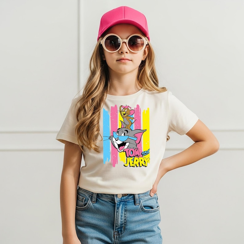 May include: A cream-coloured short-sleeve t-shirt featuring a graphic print of Tom and Jerry with blue sunglasses, set against vertical pink, blue, and yellow stripes. The shirt is paired with blue denim jeans, a bright pink baseball cap, and sunglasses.