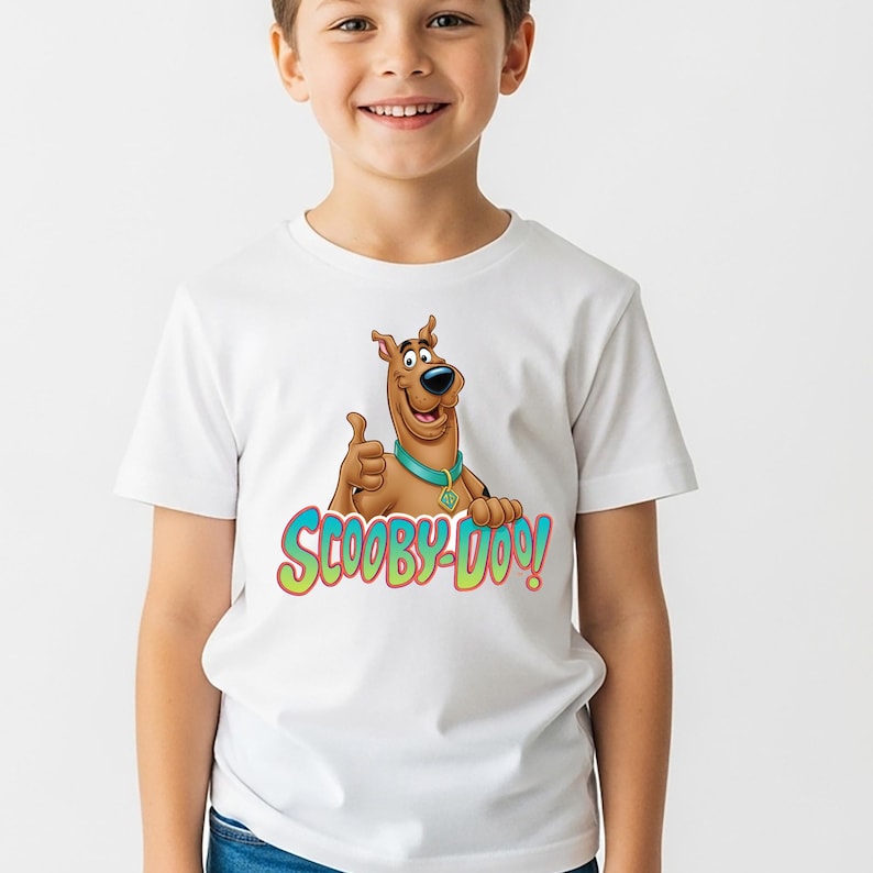 May include: A white short-sleeved t-shirt featuring a graphic of the brown cartoon dog Scooby-Doo giving a thumbs up. The character wears a teal collar with a tag. Below the character, the text SCOOBY-DOO! is printed in green and red letters.