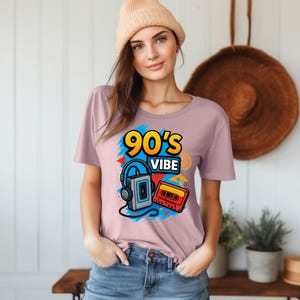 May include: A light mauve t-shirt featuring a graphic print with the text 90's VIBE in yellow and blue, accompanied by illustrations of a grey portable cassette player with black headphones and a red and yellow cassette tape on a blue background.