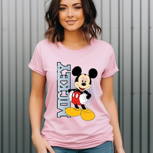 May include: A light pink short-sleeved t-shirt featuring a graphic of Mickey Mouse standing with hands on hips. The character is wearing red shorts and yellow shoes. To the left of the character, the word MICKEY is printed vertically in blue.