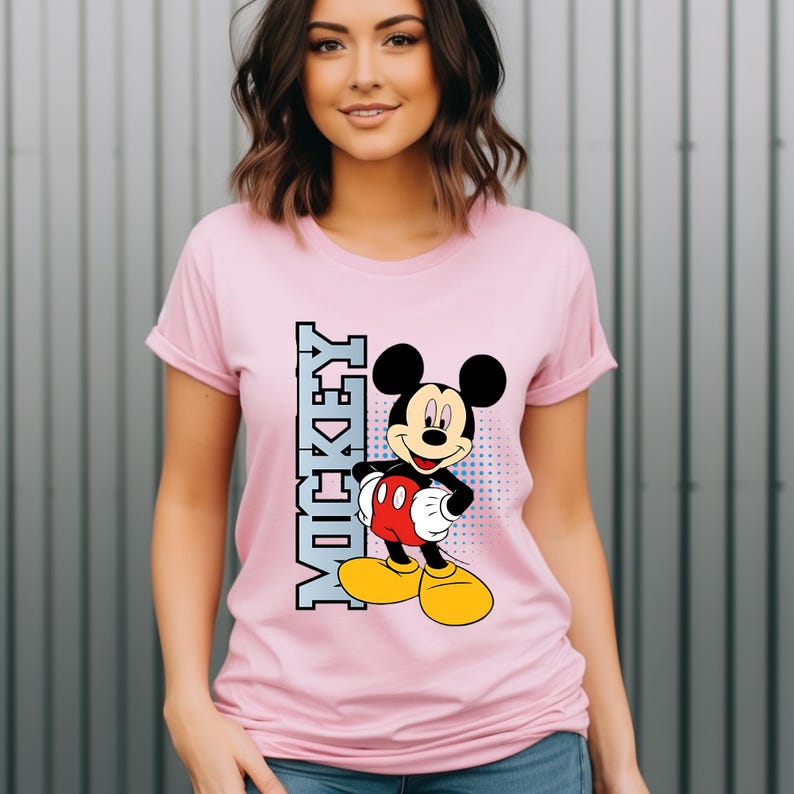 May include: A light pink short-sleeved t-shirt featuring a graphic of Mickey Mouse standing with hands on hips. The character is wearing red shorts and yellow shoes. To the left of the character, the word MICKEY is printed vertically in blue.