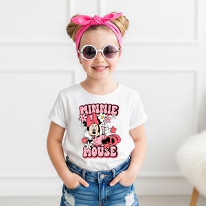 May include: A white short-sleeved t-shirt featuring a graphic print of Minnie Mouse surrounded by pink and white flowers. The text MINNIE MOUSE is printed in a bold pink font above and below the character. The shirt is paired with blue denim jeans.