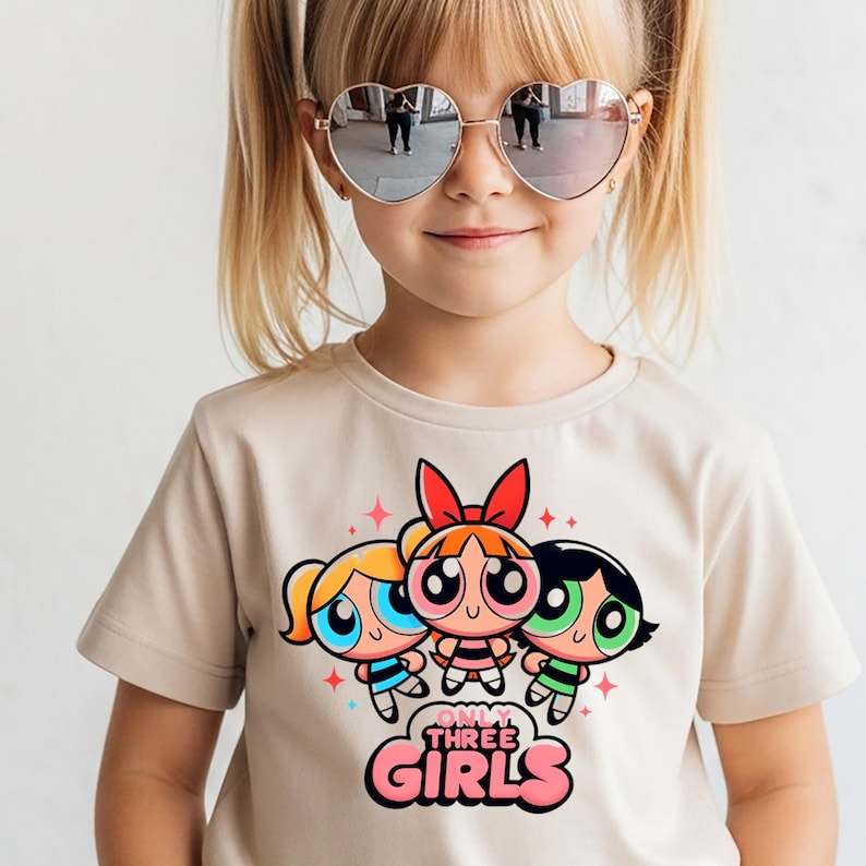May include: A beige short-sleeved t-shirt featuring a graphic print of three cartoon characters with large eyes and colourful hair. Below the characters, the text reads "ONLY THREE GIRLS" in bold, pink, rounded lettering, accented by small red star shapes.