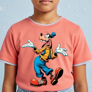 May include: A coral-coloured short-sleeved t-shirt featuring a graphic print of the cartoon character Goofy. The character is depicted with a yellow turtleneck, blue vest, blue pants, and large red shoes, standing with arms outstretched.