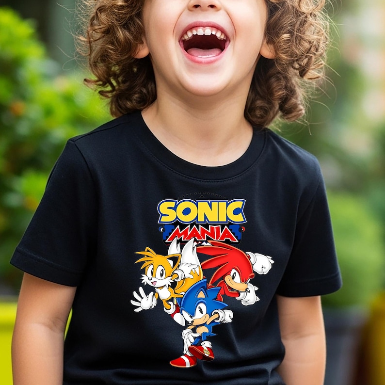 May include: A black short-sleeved t-shirt featuring a graphic print of the Sonic Mania logo in yellow and red text, positioned above colourful illustrations of the characters Sonic the Hedgehog, Tails, and Knuckles the Echidna.
