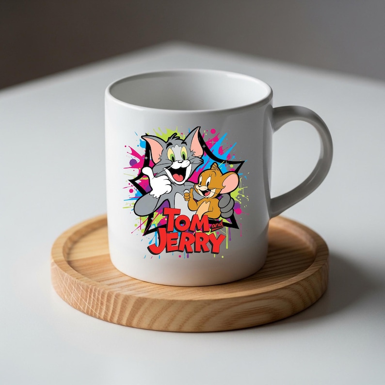 May include: A white ceramic mug featuring a colourful graphic of the cartoon characters Tom and Jerry. The design includes a black star shape, vibrant paint splatters in pink, blue, and green, and the red text Tom and Jerry printed below.