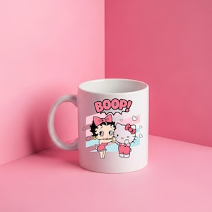 May include: A white ceramic mug featuring a graphic print of Betty Boop and Hello Kitty standing side by side. Above them, the word BOOP is printed in pink letters. The design includes small pink hearts and light blue and pink diagonal stripes.