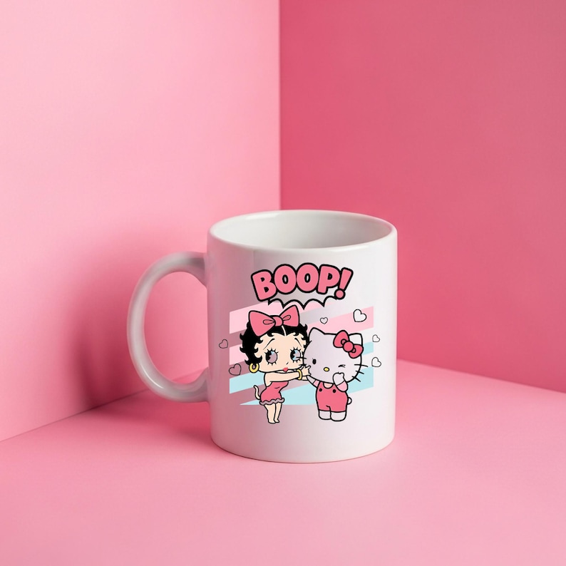 May include: A white ceramic mug featuring a graphic print of Betty Boop and Hello Kitty standing side by side. Above them, the word BOOP is printed in pink letters. The design includes small pink hearts and light blue and pink diagonal stripes.