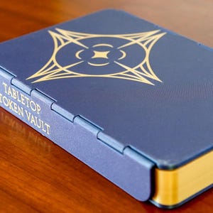 May include: A blue tabletop token vault with gold accents. The box has a gold design on the top and the words "Tabletop Token Vault" printed on the side. The edges of the box are gold.