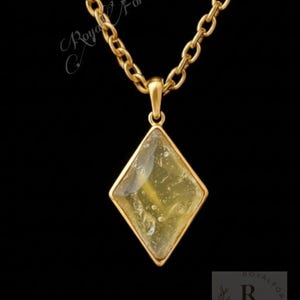 Może przedstawiać: Złoty naszyjnik z wisiorkiem w kształcie rombu. Wisiorek ma żółto-zielone, półprzezroczyste centrum i złotą ramkę. Łańcuszek ma ogniwowy wzór. Tło jest czarne.