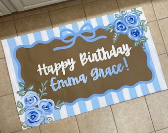 May include: A rectangular floor mat featuring a light blue and white vertical striped background. A central brown scalloped plaque displays the white text Happy Birthday Emma Grace! with a light blue bow and blue rose floral accents.
