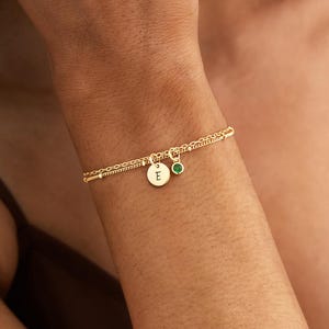 May include: A gold double-strand chain bracelet featuring a small circular charm engraved with the letter E and a separate charm with a small round green gemstone. The delicate metal links create a layered appearance on the wrist.