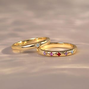 May include: Two gold rings featuring star-shaped settings. One ring has a single light blue gemstone, while the other displays a row of multicoloured gemstones including pink, red, and blue stones. The text Cherish Box Designs is visible.