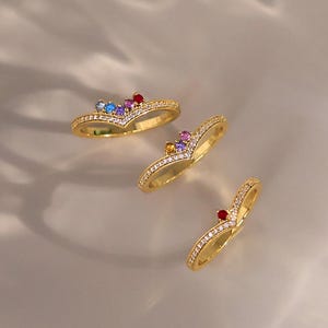May include: Three gold V-shaped rings featuring small clear pavé stones along the band. The top ring has four colored stones, the middle has three, and the bottom has one red stone. The text Cherish Box Designs is visible in the bottom right corner.