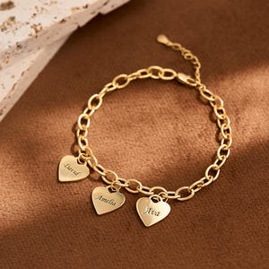 May include: Gold-coloured bracelet with a chain link design. The bracelet features three heart-shaped charms, each engraved with a name: "David", "Amelia", and "Ava". The bracelet is displayed on a brown surface.