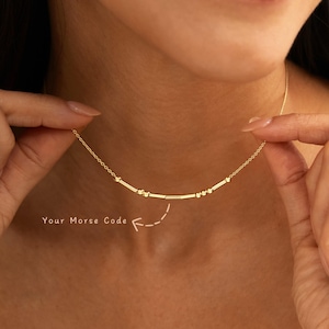 May include: A gold-toned necklace featuring a delicate chain and a central bar element composed of small beads and short rods arranged in a Morse code pattern. The text "Your Morse Code" is printed below the necklace with a dashed arrow pointing up.