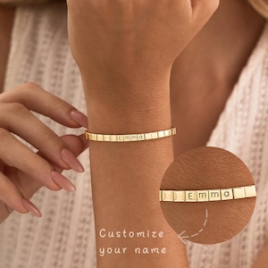 Peut inclure: Un bracelet doré composé de petits segments carrés reliés entre eux. Certains segments sont gravés avec les lettres E, M, M et A, formant le prénom Emma. Un encart circulaire montre une vue agrandie du nom avec le texte Customize your name.
