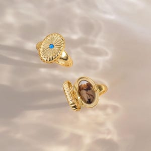 May include: Two gold rings with an oval shape. One ring features a sunburst pattern with a small blue stone in the center. The other ring is an open locket style, revealing a small photograph of two people inside the oval compartment.