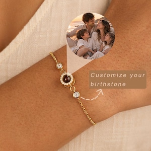 Könnte beinhalten: Ein goldfarbenes Kettenarmband mit einem zentralen runden Anhänger, der ein kleines eingebettetes Familienfoto und einen Kranz aus klaren Steinen aufweist. Zwei weitere klare Steine sind in die Kette eingesetzt. Der Text lautet: Customize your birthstone.