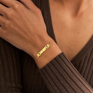 Könnte beinhalten: Ein goldfarbenes Kettenarmband mit dem Namen JENNIFER in erhabenen, abgerundeten, metallischen Buchstaben. Das Armband enthält einen kleinen, kreisförmigen, dunkelroten Edelsteinakzent, der neben dem Namensschild an einer feinen, dünnen Goldkette sitzt.