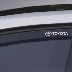 May include: A car window featuring a black trim and the white "TOYOTA" logo. The car's roof is visible in the background. The image is a close-up of the car's window.