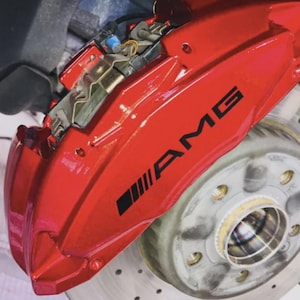 Könnte beinhalten: Nahaufnahme eines roten AMG-Bremssattels mit schwarzem AMG-Logo. Der Bremssattel ist an einer silbernen Bremsscheibe mit Bohrungen montiert. Kfz-Teile.