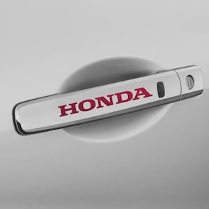 May include: A chrome car door handle with the word "HONDA" in red lettering. The handle is mounted on a white car door, with a subtle reflection visible. The handle has a sleek, modern design.