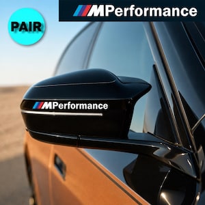 Op de afbeelding: Een glanzend zwarte autospiegelkap voorzien van het MPerformance-logo in witte letters met een blauwe, paarse en rode streepafbeelding. De sticker is aangebracht op de buitenbehuizing van de spiegel die op een autodeur is gemonteerd.