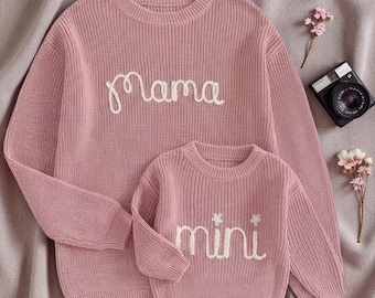 May include: Matching pink sweaters, one adult and one child size, with the words "Mama" and "Mini" embroidered in white script. The sweaters are made of a ribbed knit fabric.