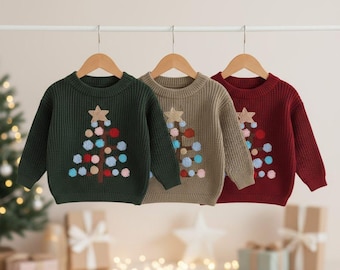 May include: Three children's sweaters in green, tan, and burgundy, each with a Christmas tree design. The tree is embroidered with a star and colorful pom-poms. The sweaters are hanging on wooden hangers.