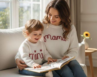 May include: A mother and child are seated on a white sofa, reading a book together. Both are wearing matching cream-colored sweaters. The mother's sweater says "mama" in burgundy script, and the child's says "mini".