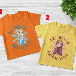 May include: Two short-sleeved t-shirts. One is orange with a graphic of Elsa and the text "Big Sister I'm her Elsa". The other is yellow with Anna and the text "Lil' Sister I'm her Anna".