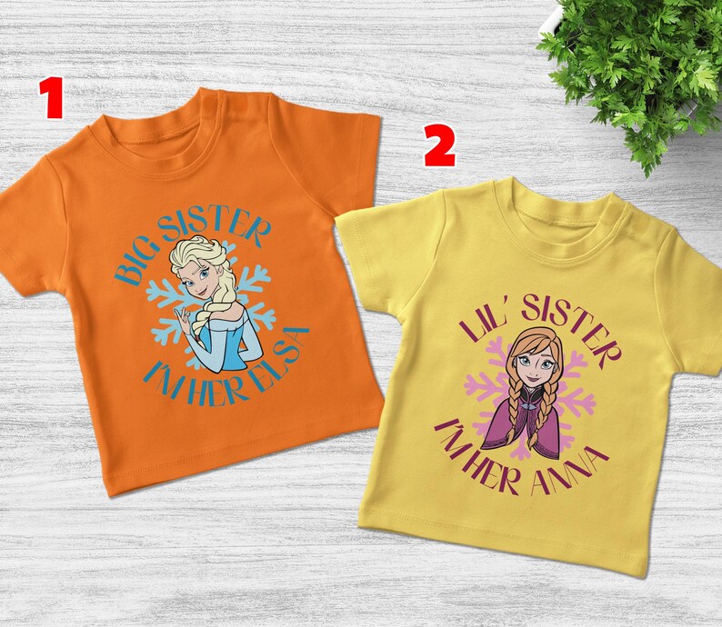 May include: Two short-sleeved t-shirts. One is orange with a graphic of Elsa and the text "Big Sister I'm her Elsa". The other is yellow with Anna and the text "Lil' Sister I'm her Anna".