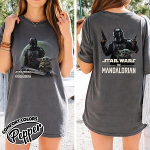 May include: Dark gray t-shirt featuring a graphic of The Mandalorian and Grogu from Star Wars. The front graphic shows the characters with the text "STAR WARS THE MANDALORIAN". The back graphic shows the Mandalorian holding blasters with Grogu and the same text.