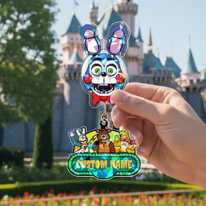 May include: A clear acrylic keychain featuring a cartoon bunny character with blue ears, a red bow tie, and green eyes. Below, a group of cartoon characters are displayed above a green and blue sign that reads "CUSTOM NAME".