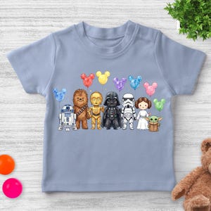 Op de afbeelding: Een lichtblauw T-shirt met een kleurrijke illustratie van Star Wars-personages die elk een Mickey Mouse-vormige ballon vasthouden. De personages zijn R2-D2, Chewbacca, C-3PO, Darth Vader, een Stormtrooper, prinses Leia en Grogu.