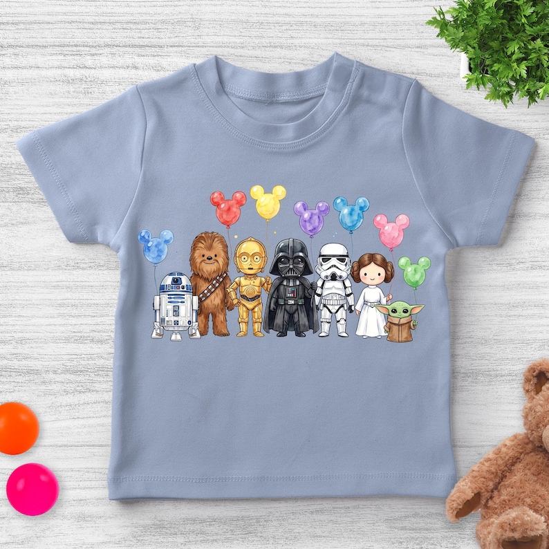 Op de afbeelding: Een lichtblauw T-shirt met een kleurrijke illustratie van Star Wars-personages die elk een Mickey Mouse-vormige ballon vasthouden. De personages zijn R2-D2, Chewbacca, C-3PO, Darth Vader, een Stormtrooper, prinses Leia en Grogu.