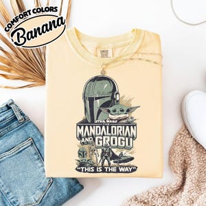 Puede incluir: Una camiseta amarilla clara con un gr&aacute;fico de Star Wars de The Mandalorian y Grogu. El dise&ntilde;o incluye el casco del Mandaloriano, Grogu y el texto "THIS IS THE WAY". La camiseta tambi&eacute;n tiene el texto "COMFORT COLORS" y "Banana".
