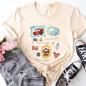 Puede incluir: Camiseta beige con gr&aacute;ficos de coches de dibujos animados coloridos. El dise&ntilde;o incluye im&aacute;genes enmarcadas de coches, una bandera a cuadros y un globo. La camiseta se combina con vaqueros grises y zapatillas negras.