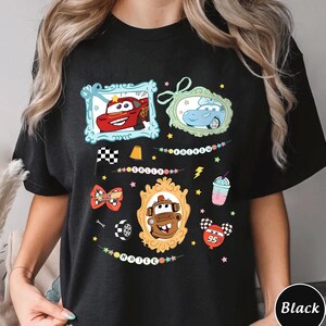 Puede incluir: Camiseta negra con personajes de coches de dibujos animados en marcos decorativos. El dise&ntilde;o incluye un coche rojo, un coche azul y un coche marr&oacute;n, adem&aacute;s de estrellas, una bandera a cuadros y un batido.